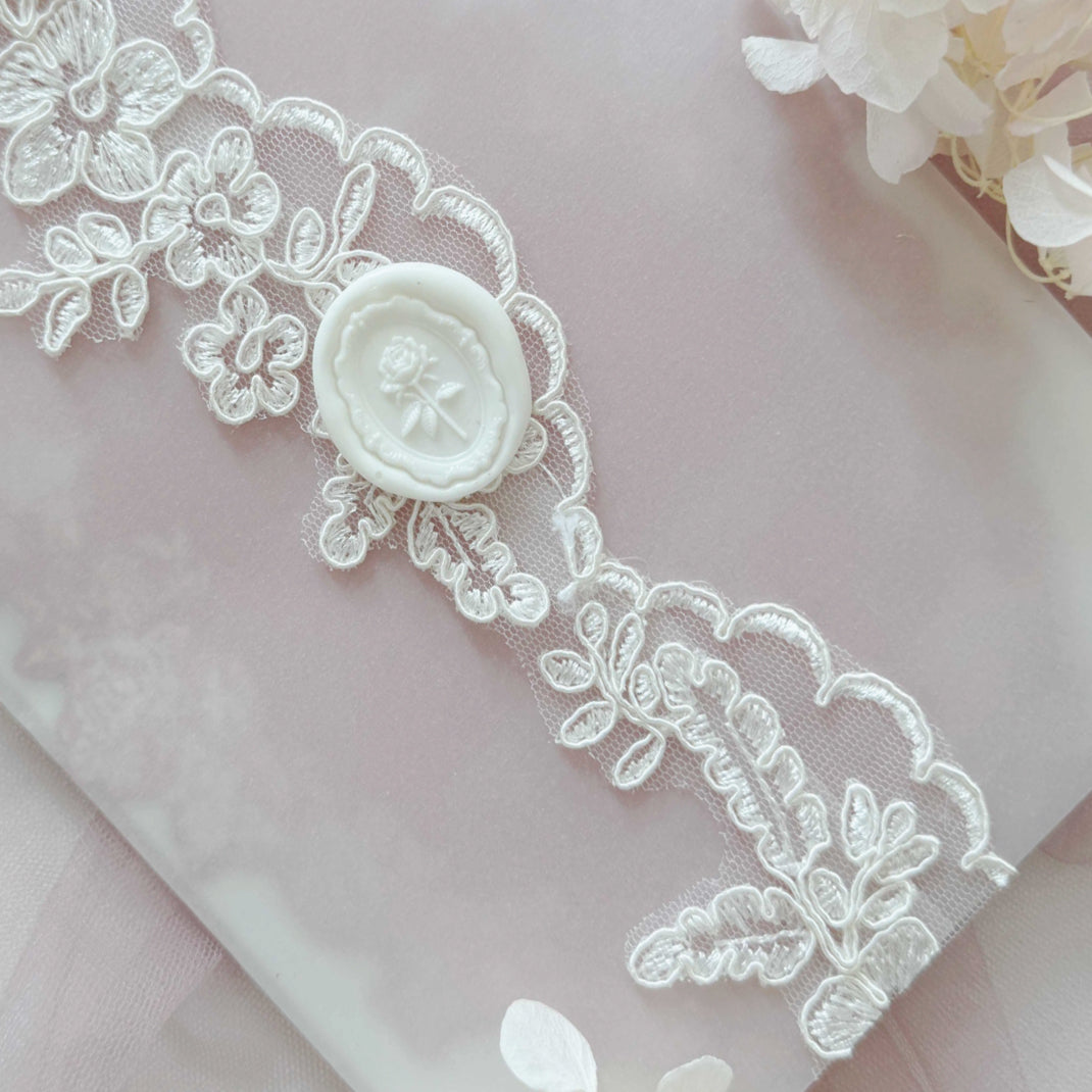 Close-up of white lace wedding invitation card with floral details on a light pink fabric background.