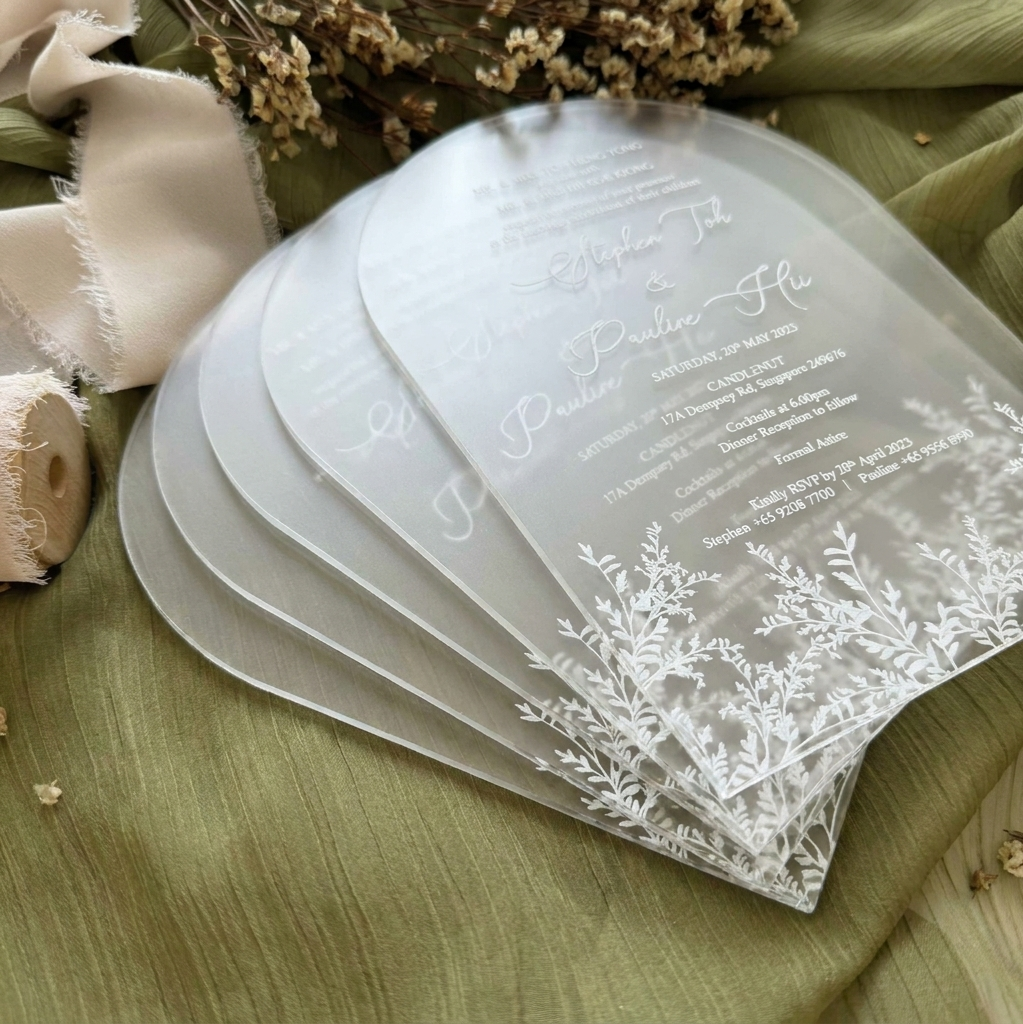 Stack of transparent wedding invitations with floral designs on a green fabric background